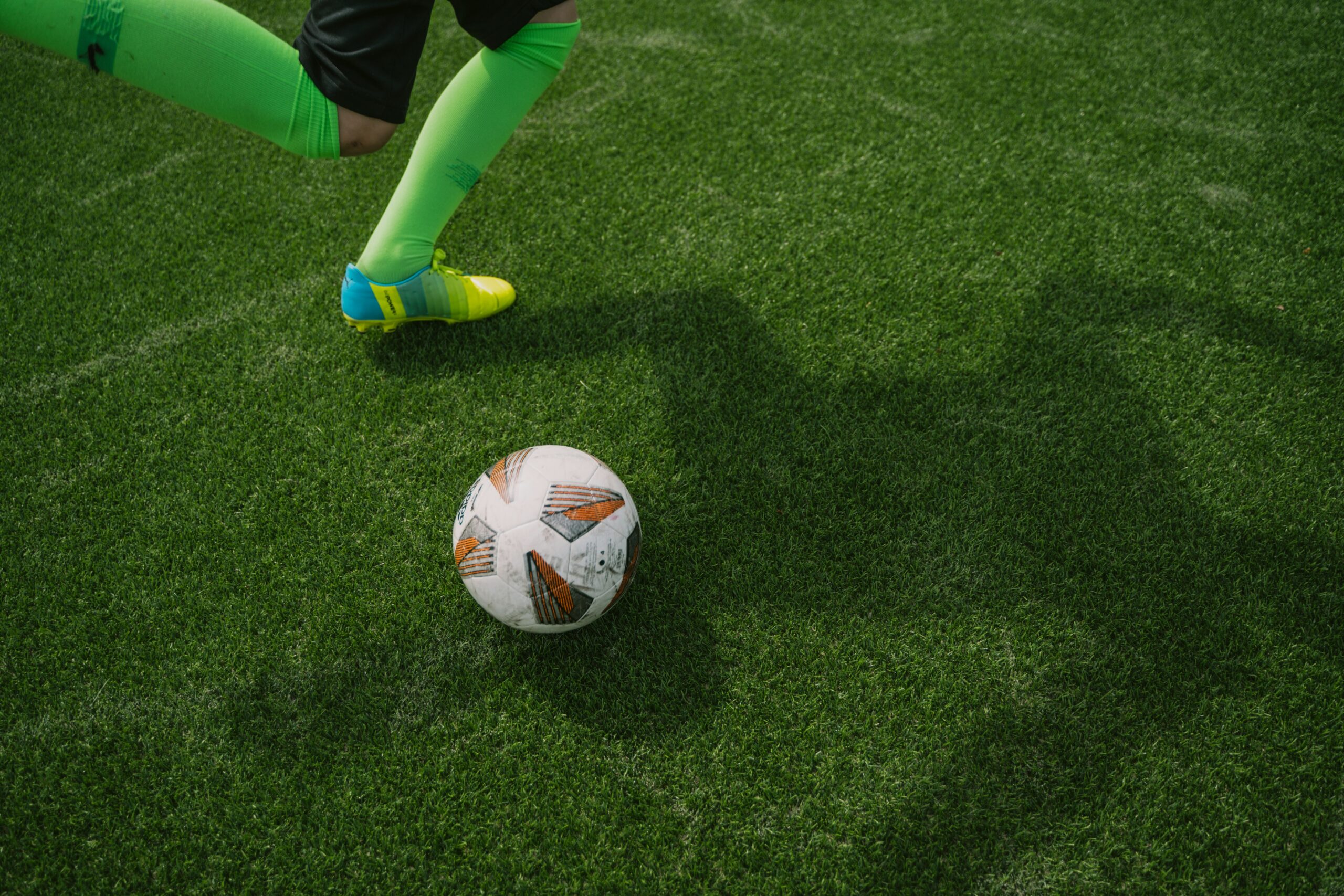Ein Kind beim Fußballtraining auf dem Kunstrasen; kurz vor dem Schuss auf den Ball, mit grünen Stutzen und bunten Fußballschuhen.